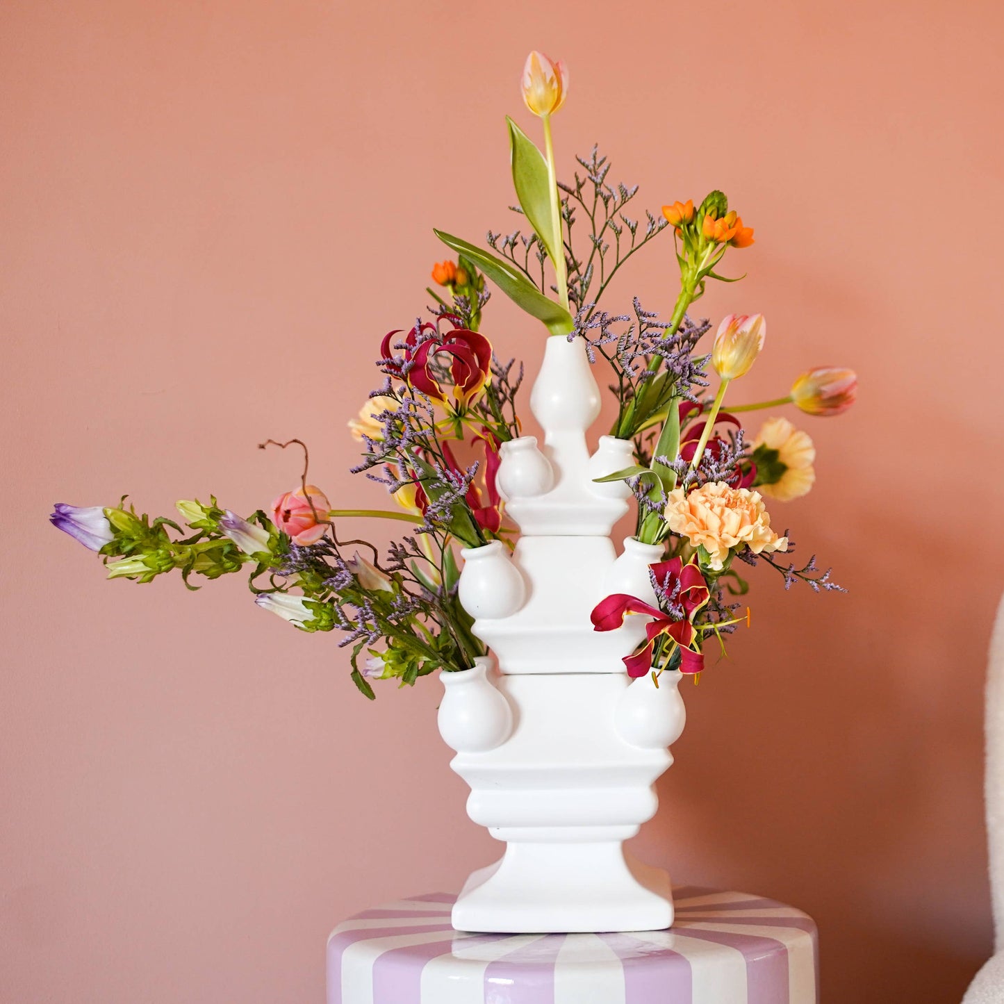 White vase with colorful flowers on a table next to a sofa with a pink and yellow pillow against a peach wall.
