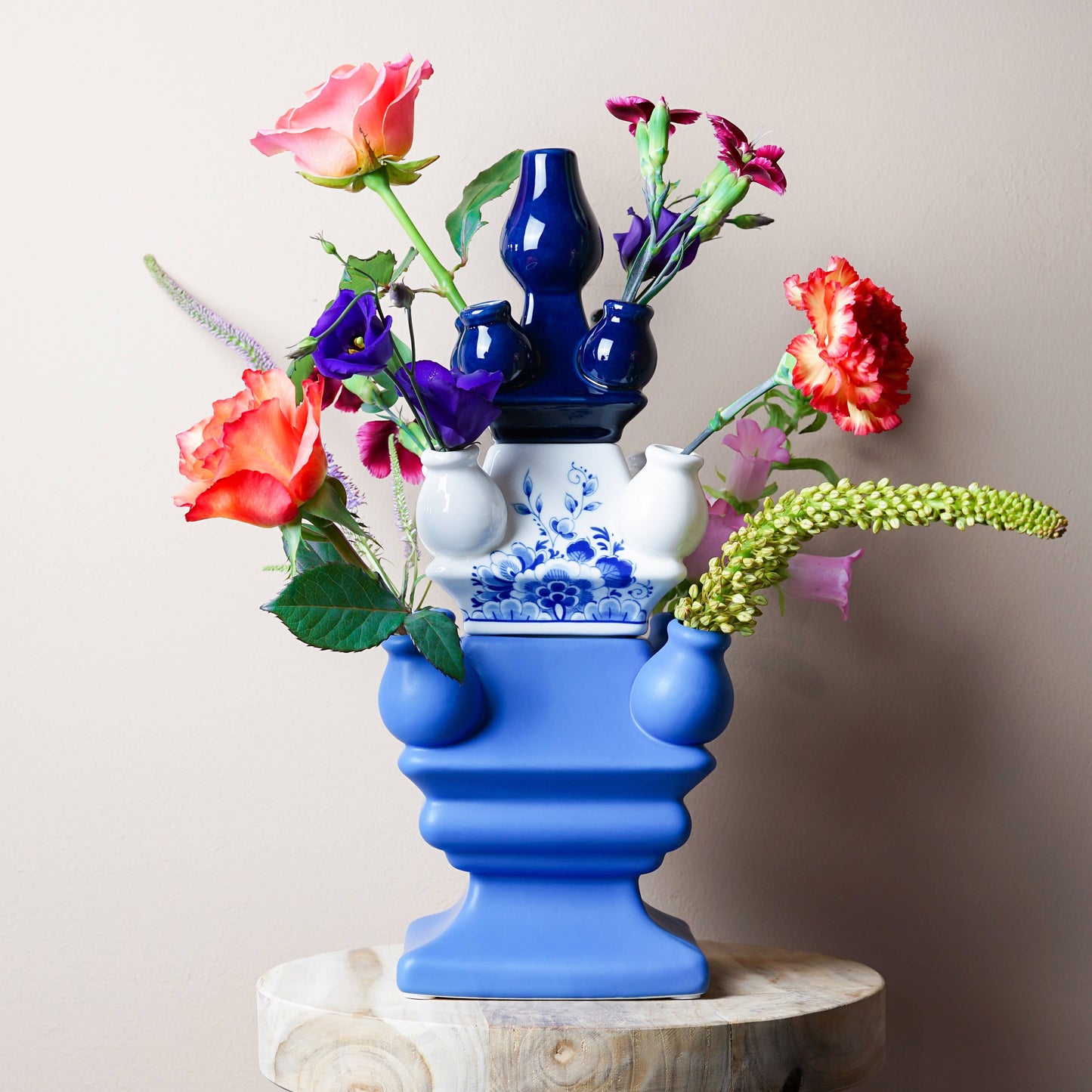Colorful floral arrangement in a blue and white vase on a wooden stool against a plain background